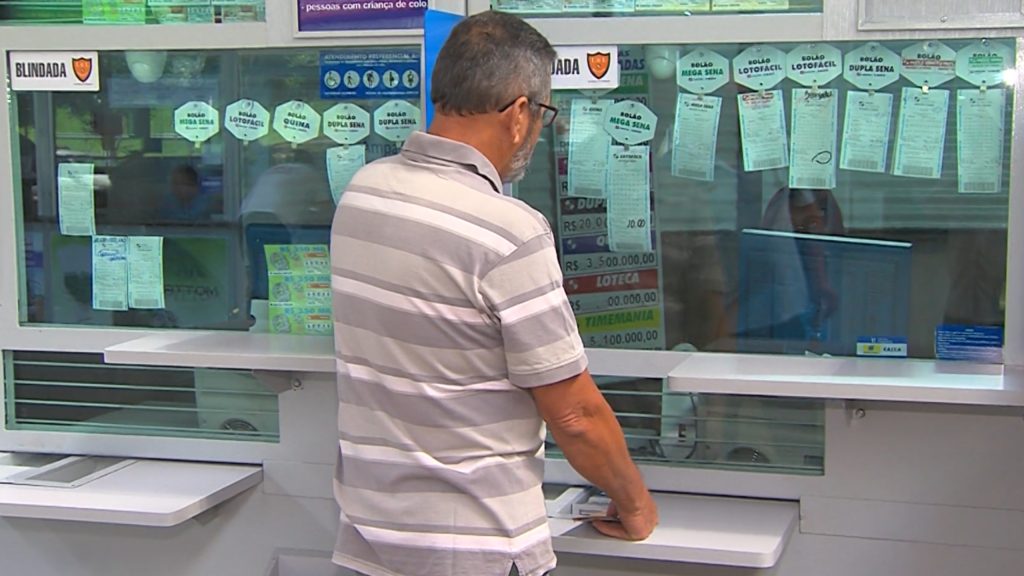 Homem de blusa listrada em frente a um caixa da lotérica pagando o seu jogo na lotomania