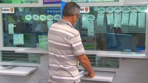 Homem de blusa listrada em frente a um caixa da lotérica pagando o seu jogo na mega-sena