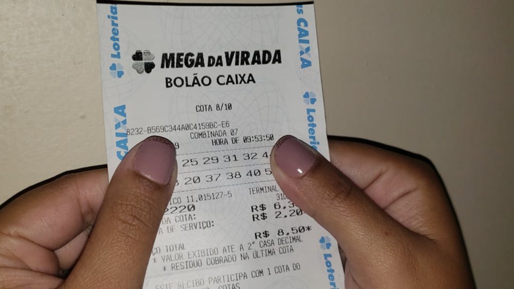 Imagem da mão de uma mulher com as unhas pintadas segurando um jogo da mega da virada que ela fez pela lotérica