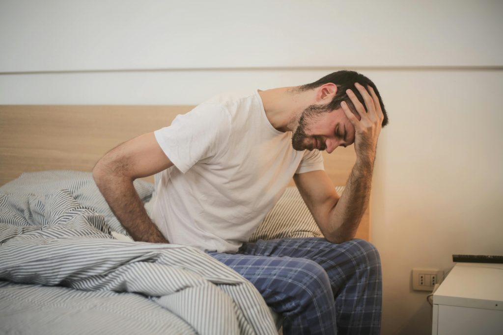 Homem sentado na cama com a mão na cabeça, parecendo não estar se sentindo bem