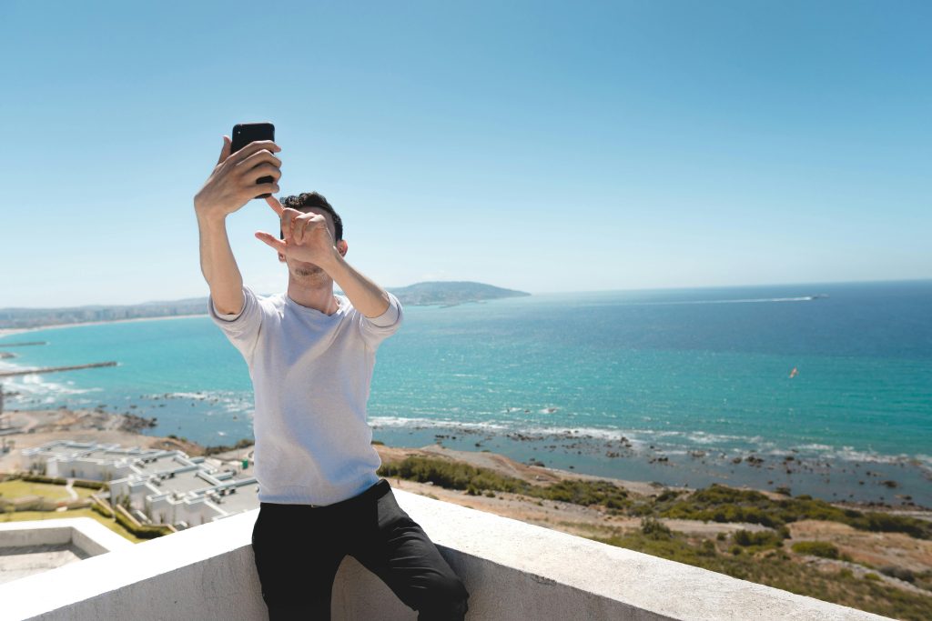 Homem sentado na beirada de um muro para tirar uma selfie com a paisagem