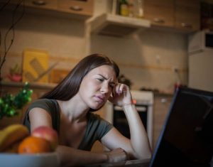 Mulher sentada em uma mesa, de frente para um notebook, com feição de dor