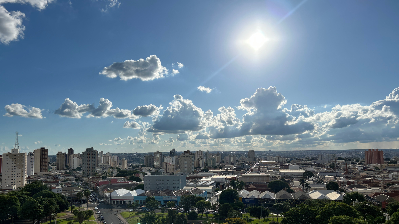 Cel em Uberlândia azul, com nuvens e sol ao centro. Abaixo está a cidade com muitos prédios, comércios e residências