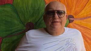Cássio Teodoro com óculos escuro e uma parede com imagens de flores ao fundo