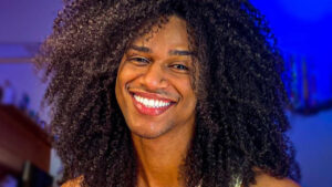Murilo Lorran sorrindo, em um comodo de uma residência e luz azul ao fundo