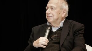 Luis Fernando Verissimo olhando para o lado durante entrevista. Vestido com terno preto e de microfone na mão.