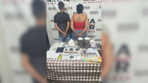 Foto da PM com o casal preso de costas, algemados e à frente os materias apreendidos encontrados do depósito de drogas e no carro do casal