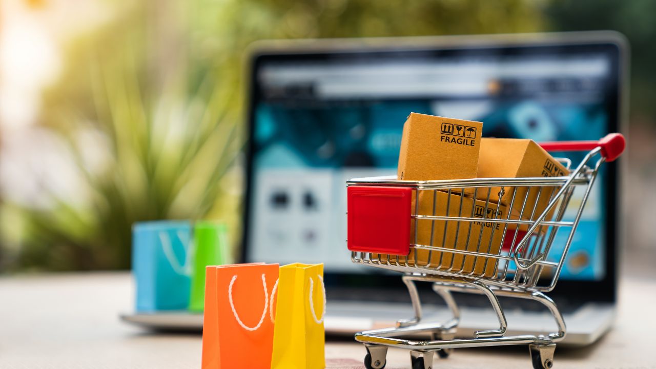 carrinho de compras cheio com um computador desfocado ao fundo representando compras online