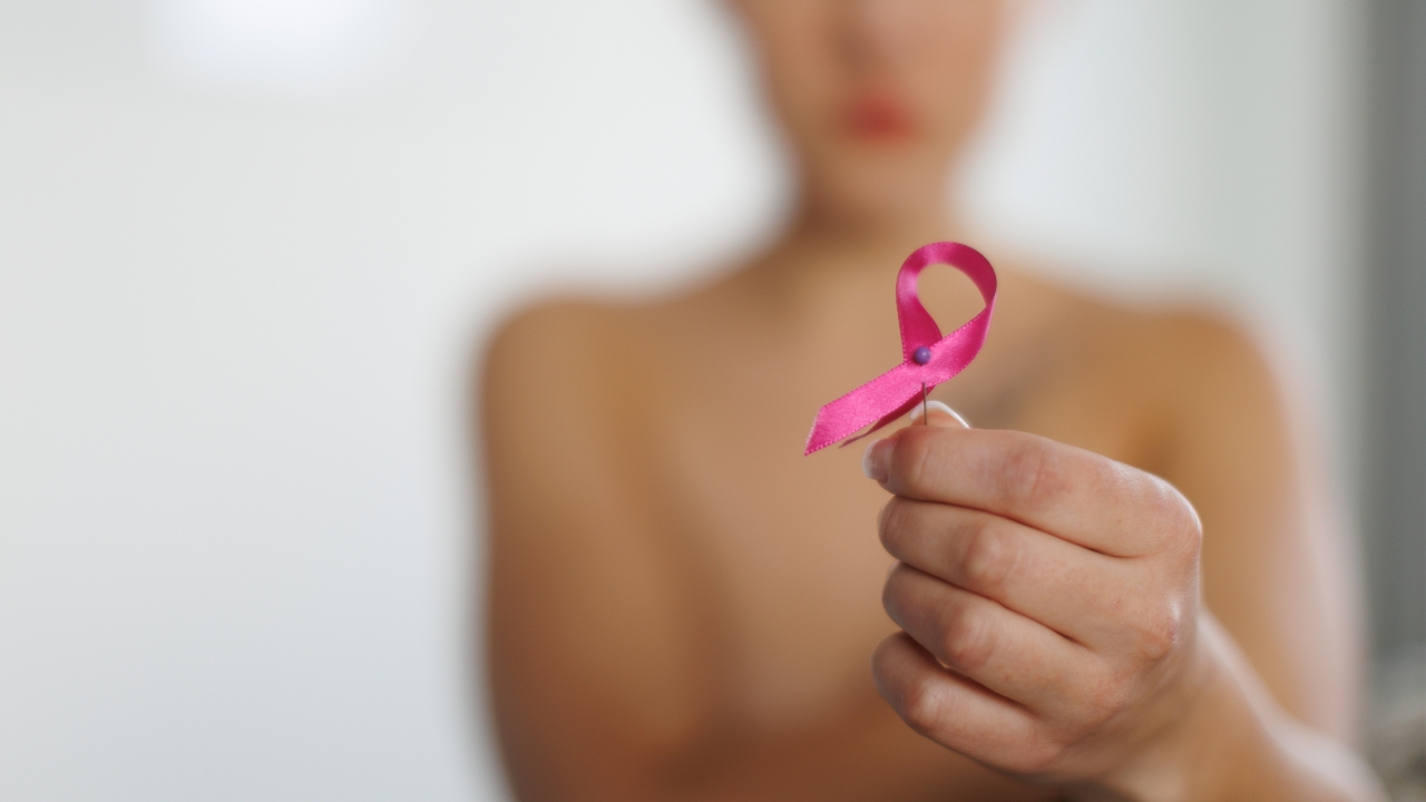 Mulher desfocada ao fundo, sefurando o laço símbolo do Outubro Rosa