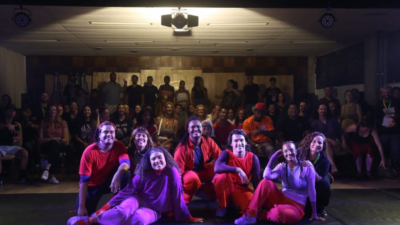 Grupo de pessoas sentadas em primeiro plano com plateia ao fundo Grupo de pessoas sentadas em primeiro plano com plateia ao fundo